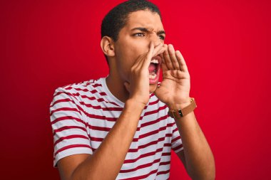 Genç yakışıklı arap adam izole kırmızı arka plan üzerinde çizgili t-shirt giyen ağız üzerinde elleri ile yüksek sesle kızgın bağırarak