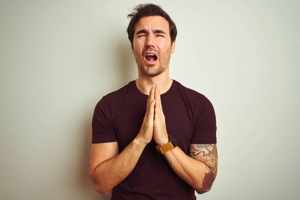 Young handsome man with tattoo wearing purple casual t-shirt over isolated white background begging and praying with hands together with hope expression on face very emotional and worried. Asking for forgiveness. Religion concept.