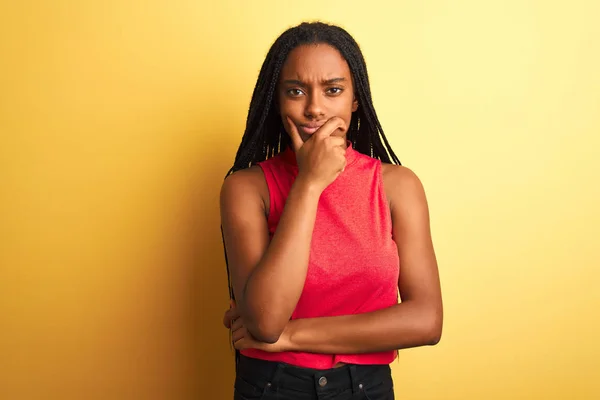 African american woman wearing red casual t-shirt standing over isolated yellow background looking confident at the camera with smile with crossed arms and hand raised on chin. Thinking positive.