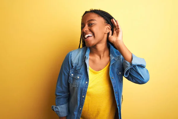 Young african american woman wearing denim shirt standing over isolated yellow background smiling with hand over ear listening an hearing to rumor or gossip. Deafness concept.