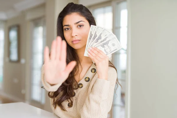 Young woman holding 20 dollars bank notes with open hand doing stop sign with serious and confident expression, defense gesture