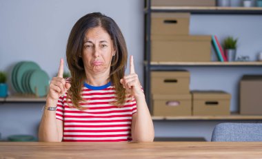 Orta yaş yaşlı kadın evde masada oturan üzgün ve üzgün görünümlü kadar işaret, parmakları ile yön gösteren, mutsuz ve depresif.