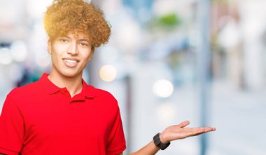 Neşeli sunulması ve kameraya bakarak avucunun ile işaret gülümseyen kırmızı t-shirt giyen afro saçlı Genç yakışıklı adam.