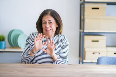 Orta yaş yaşlı kadın evde masada oturan tiksinti ifade, hoşnutsuz ve korkulu tiksinti yüz yapıyor çünkü nefret tepki. Elleri havada. Sinir bozucu kavram.