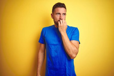 Genç yakışıklı adam sarı izole arka plan üzerinde rahat mavi t-shirt giyen stresli ve ağız tırnak ısırma elleri ile sinir görünümlü. Anksiyete sorunu.