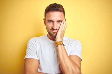 Genç yakışıklı adam sarı izole arka plan üzerinde rahat beyaz t-shirt giyen yorgun görünümlü ve çapraz kolları ile depresyon sorunları ile sıkılmış düşünme.