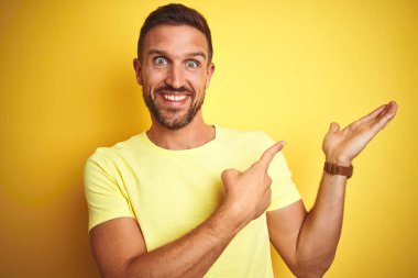 Genç yakışıklı adam sarı izole arka plan üzerinde rahat sarı t-shirt giyen hayran ve el ile sunarken kameraya gülümseyerek ve parmak ile işaret.