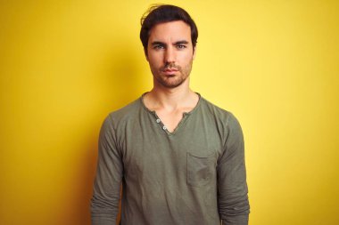 Izole sarı arka plan üzerinde duran rahat t-shirt giyen genç yakışıklı adam yüzünde ciddi ifade ile rahat. Kameraya basit ve doğal bir bakış.