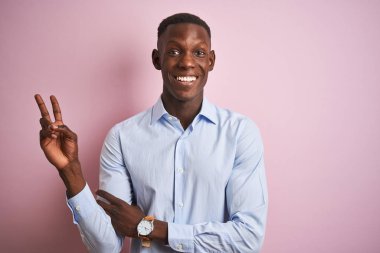 Mavi zarif gömlek giyen Afro-amerikan adam izole pembe arka plan üzerinde duran mutlu yüz ile gülümseyerek zafer işareti yapıyor kameraya göz kırparak. İki numara.