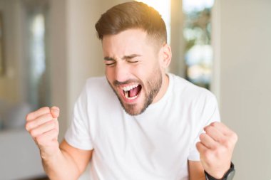 Evde rahat beyaz t-shirt giyen genç yakışıklı adam çok mutlu ve heyecanlı silah kaldırdı, gülümseyen ve başarı için çığlık ile kazanan jest yapıyor. Kutlama konsepti.