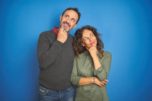 Beautiful middle age couple together standing over isolated blue background with hand on chin thinking about question, pensive expression. Smiling with thoughtful face. Doubt concept.