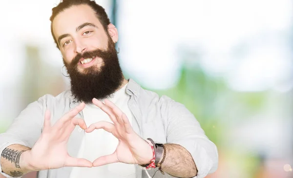 Young man with long hair, beard and earrings smiling in love showing heart symbol and shape with hands. Romantic concept.