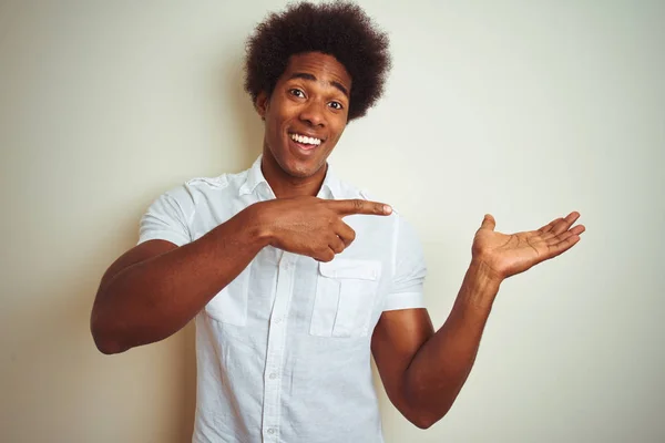 African american man with afro hair wearing shirt standing over isolated white background amazed and smiling to the camera while presenting with hand and pointing with finger.