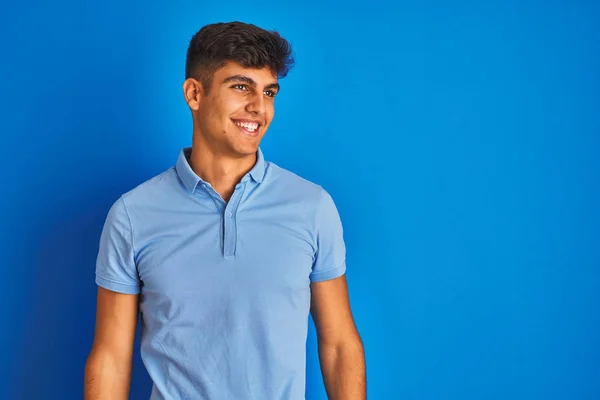 カジュアルなポロを着た若いインド人男性が 青い背景の上に立ち 笑顔で横を向き 自然な表情 自信を持って笑う — ストック写真
