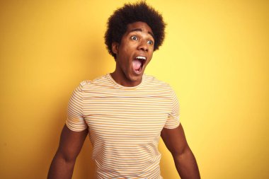 Afro saç ile Amerikan adam izole sarı arka plan üzerinde duran çizgili t-shirt ayakta kızgın ve deli çığlık sinirli ve öfkeli, öfke ile bağırıyor. Öfke ve agresif kavram.