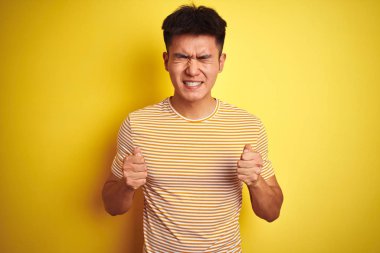 İzole sarı arka plan üzerinde duran tişört giyen genç Asyalı çinli adam, kollarını kaldırıp gözleri kapalı zaferi gülümseyerek başarı için heyecanlı. Kazanan kavramı.