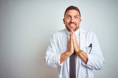 İzole arka plan üzerinde beyaz profressional ceket giyen genç yakışıklı doktor adam birlikte emin gülümseyerek af isteyen elleriyle dua.