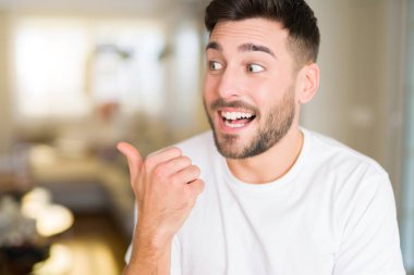 Evde rahat beyaz t-shirt giyen genç yakışıklı adam arıyor ve başparmak yanında işaret mutlu yüz ile gülümseyen.