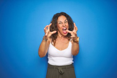 Kıvırcık saçlı orta yaş yaşlı kadın mavi izole arka plan üzerinde duran öfke ile sinirli bağırarak, elleri boğmaya çalışıyor, deli bağırıyor