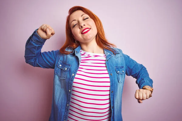 Beautiful redhead woman wearing denim shirt and striped t-shirt over isolated pink background stretching back, tired and relaxed, sleepy and yawning for early morning
