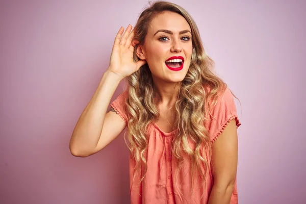年轻的漂亮女人穿着T恤站在粉红色的孤立背景微笑与手在耳朵听听传闻或八卦 耳聋概念 — 图库照片
