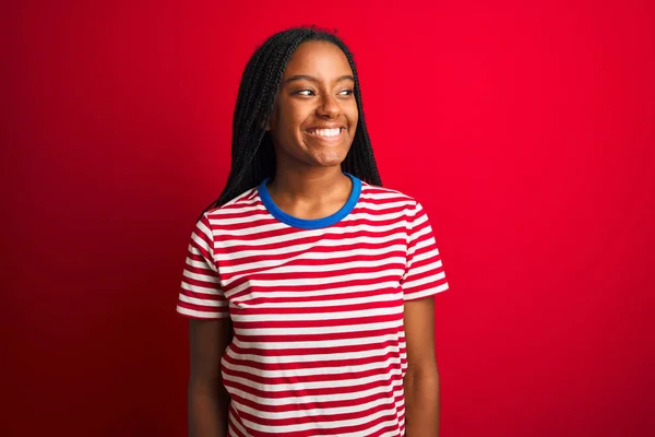縞模様のTシャツを着た若いアフリカ系アメリカ人女性は 顔に笑顔で 自然な表情で目を背けている孤立した赤い背景の上に立っている 自信を持って — ストック写真