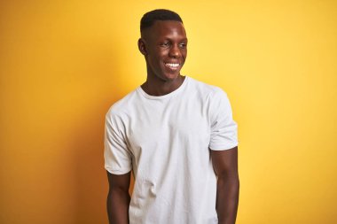 Beyaz tişört giyen genç afro-amerikan adam izole sarı arka plan üzerinde duran yüzünde gülümseme, doğal ifade ile yan bakıyor. Gülmekten emin.