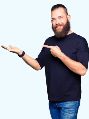 Genç Sarışın giyiyor rahat t-shirt hayrete ve el ile sunulması ve parmak ile işaret ederken kameraya gülümseyen adam.