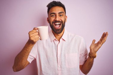 Genç hintli adam izole pembe arka plan üzerinde duran kahve fincan içme giyen çok mutlu ve heyecanlı, kazanan ifade büyük bir gülümseme ile çığlık zafer kutluyor ve ellerini kaldırdı
