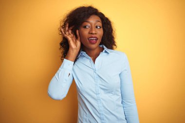 Izole sarı arka plan üzerinde zarif gömlek giyen Afro-Amerikan iş kadını el kulak üzerinden bir söylenti veya dedikodu bir işitme dinlerken gülümseyerek. Sağırlık kavramı.