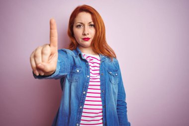 Denim gömlek giyen güzel kızıl saçlı kadın ve izole pembe arka plan üzerinde çizgili t-shirt parmak yukarı ve kızgın ifade ile işaret, hiçbir jest gösteren