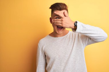 Beyaz rahat t-shirt giyen genç yakışıklı adam şok el ile yüz ve gözleri kapsayan şok gözetleme izole sarı arka plan üzerinde duran, utanmış ifade ile parmaklarından bakarak.