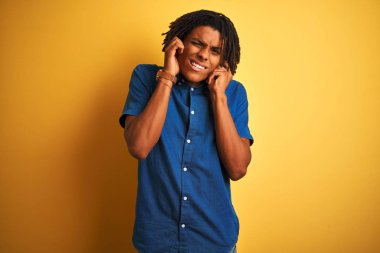 Dreadlocks ile rahat kot gömlek giyen Afro adam yüksek sesle müzik sesi için rahatsız ifade ile parmakları ile kulakları kapsayan izole sarı arka plan üzerinde duran. Sağır kavramı.