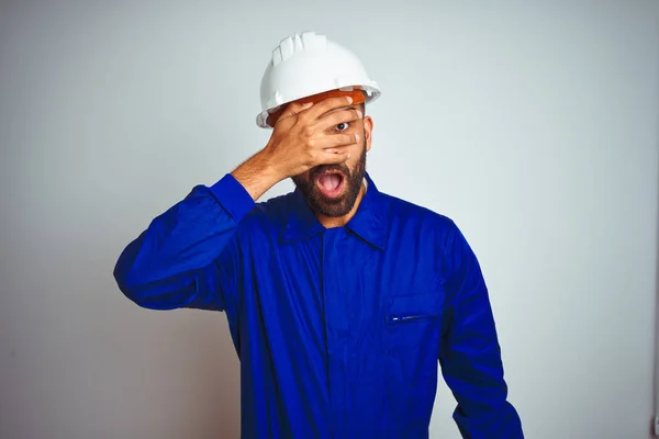 Handsome indian worker man wearing uniform and helmet over isolated white background peeking in shock covering face and eyes with hand, looking through fingers with embarrassed expression.