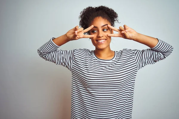 African american woman wearing navy striped t-shirt standing over isolated white background Doing peace symbol with fingers over face, smiling cheerful showing victory