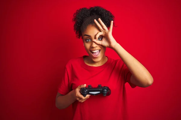 African american gamer woman playing video game using joystick over isolated red background with happy face smiling doing ok sign with hand on eye looking through fingers