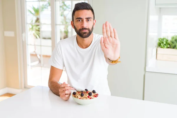 Handsome hispanic man eating healthy breakfast in the morning at home with open hand doing stop sign with serious and confident expression, defense gesture