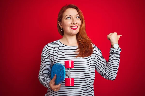 Genç kızıl saçlı öğrenci kadın kırmızı izole arka plan üzerinde paten tutan işaret ve mutlu yüzü gülümseyerek yan başparmak ile gösteren