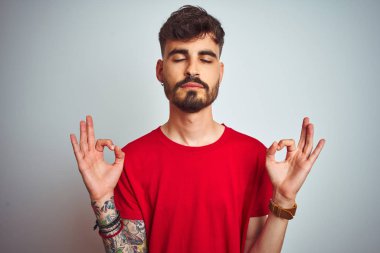 Kırmızı t-shirt giyen dövme giyen genç adam izole beyaz arka plan üzerinde duran rahatlayın ve gözleri parmakları ile meditasyon jest yapıyor kapalı gülümseyerek. Yoga konsepti.