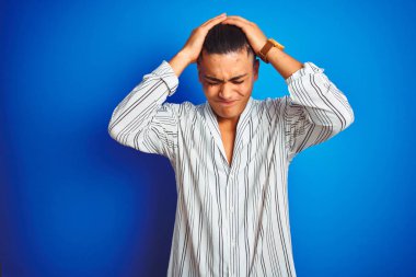 Çizgili gömlek giyen genç brezilyalı adam izole mavi arka plan üzerinde ayakta baş ağrısı umutsuz muzdarip ve stresli çünkü ağrı ve migren. Eller baş üstünde.