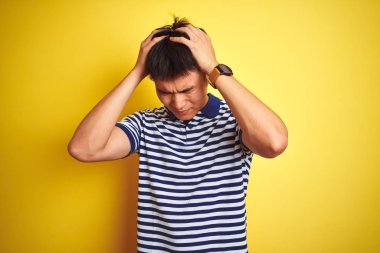 Genç Asyalı çinli adam çizgili polo giyen izole sarı arka plan üzerinde ayakta baş ağrısı umutsuz muzdarip ve stresli çünkü ağrı ve migren. Eller baş üstünde.