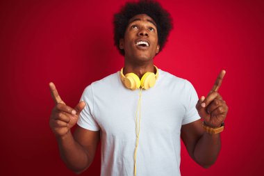 Genç afro-Amerikan adam izole kırmızı arka plan üzerinde kulaklık kullanarak müzik dinlerken şaşırttı ve baktı ve parmakları ile işaret ve kolları kaldırdı şaşırttı.