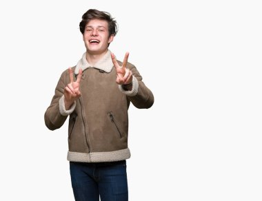 İzole arka plan üzerinde kış lık palto giyen genç yakışıklı adam gülümseyerek kameraya bakarak parmaklarını zafer işareti yaparken gösteren. İki numara.