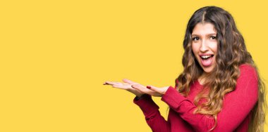 Kırmızı kazak giyen genç güzel kadın el ve açık avuç içi ile yan işaret, mutlu ve kendinden emin gülümseyen reklam sunan