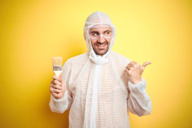 Genç adam ressam ekipman giyen ve izole sarı arka plan üzerinde boyama fırçası tutan işaret ve mutlu yüzü gülümseyerek yan başparmak ile gösteren