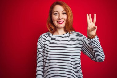 Genç kızıl saçlı kadın kırmızı izole arka plan üzerinde duran askılı lacivert gömlek giyen gösteren ve emin ve mutlu gülümserken parmakları ile üç işaret.