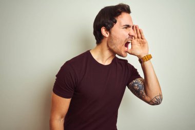 Dövme ile genç yakışıklı adam izole beyaz arka plan üzerinde mor rahat t-shirt giyen bağırarak ve ağızda el ile yan yüksek sesle bağırıyor. İletişim kavramı.
