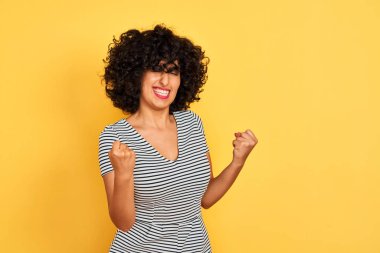 İzole sarı arka plan üzerinde çizgili elbise giyen kıvırcık saçlı genç Arap kadın çok mutlu ve heyecanlı kolları kaldırdı ile kazanan jest yapıyor, gülümseyerek ve başarı için çığlık. Kutlama konsepti.