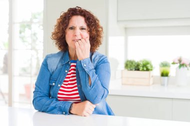 Kıvırcık saçlı orta yaş yaşlı kadın evde kot ceket giyen stresli ve ağız tırnak ısırma elleri ile sinirli görünümlü. Anksiyete sorunu.