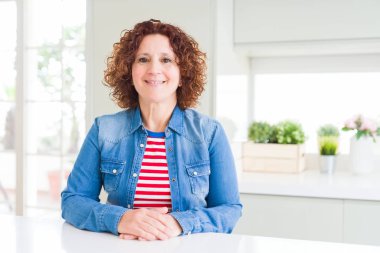 Kıvırcık saçlı orta yaş yaşlı kadın yüzünde mutlu ve serin bir gülümseme ile evde kot ceket giyiyor. Şanslı kişi.